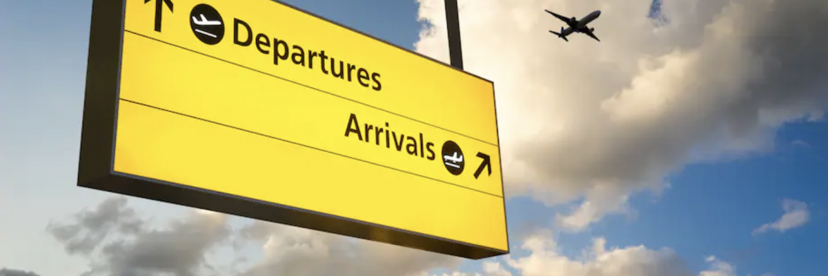 In this image is a hanging departure and arrival sign outside an airport. An airplane is flying in the sky in the background. (iStock)