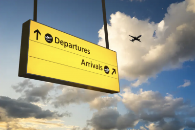 In this image is a hanging departure and arrival sign outside an airport. An airplane is flying in the sky in the background. (iStock)